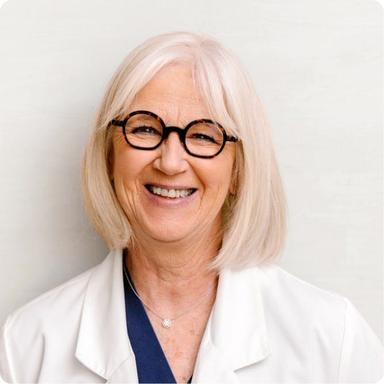 Portrait of a smiling older female healthcare provider with shoulder-length white hair, round black glasses, and a white coat.